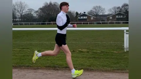 Keele University Spencer is running on a brown track with grass in the background. He is wearing a long white top and black shorts, with yellow running shoes. He has his right arm strapped onto his chest in a black bandage,