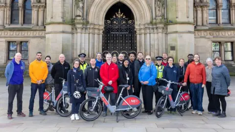 LDRS About 25 people stand outside Bradford City Hall with three red and grey e-bikes