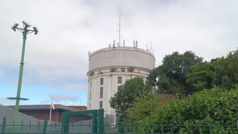 BBC A round, white water tower set in grassland but surrounded by other water works buildings, trees and green metal security fencing.