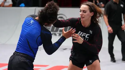 Getty Images Ffion Davies fighting Cassia Moura. She has her hand on Moura's head attempting a grappling move. She is wearing a black UFC BJJ branded top.