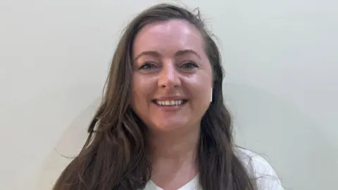 Sarah Dugdale Sarah Dugdale is standing in front of a white wall. She has long brown hair and is wearing a white T-shirt.