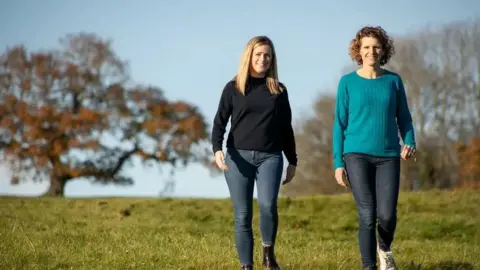 Women's Work Lab Camilla Rigby and Rachel Mostyn are walking next to each other down a grass hill. There are autumnal trees in the background. They are both wearing jeans and jumpers.