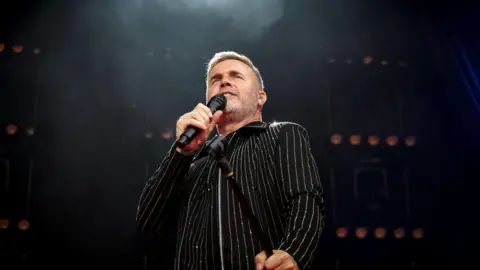Cuffe and Taylor Gary Barlow, wearing a black sparkly shirt, sings into a microphone on stage
