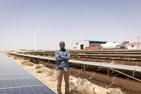 Alyona Synenko/ICRC One of the largest electricity and energy providers in Somalia, NECSOM is a hybrid fossil fuel and green energy producer. Here a smiling man is seen standing in front of banks of solar panels, arms folded across his chest.