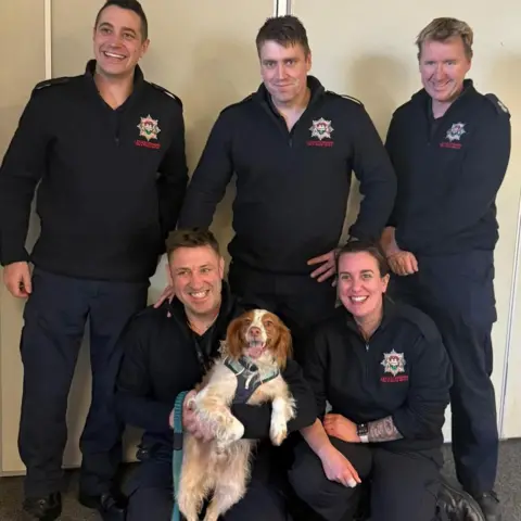 Leicestershire Fire and Rescue Service Five firefighters grinning in uniform as they cuddle up with a dog. Three of them, men, are standing, while a man and a woman kneels. The kneeling man holds a dog with brown ears and brown/white fur on the rest of its body with a green harness around its neck and torso.