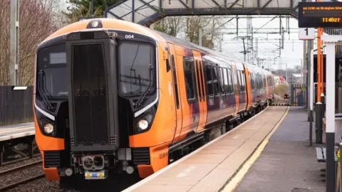 A West Midlands Railways train at a station platform. It has three carriages and is orange and purple. There is a bridge that goes over the railway lines from one platform to the other opposite