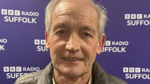 George Burley is smiling and looking directly at the camera. He is wearing a shirt and body warmer. He is standing in front of a BBC Suffolk purple background.
