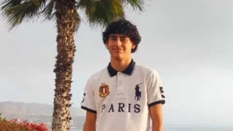 GoFundMe A teenage boy with dark curly hair and wearing a white t-shirt. He is standing at a beach. There is a palm tree behind him.