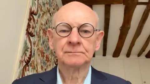 A man sat down looking at the computer camera wearing circular glasses, a navy suit with a light blue collared shirt underneath.