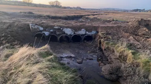 Five newly installed large concrete pipes sit under a footpath which passes over some tidal mudflats. The construction allows water to pass under the raised path.