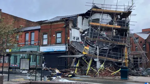 Mat Trewern/BBC Debris and a smashed bus stop are in front of a partially collapsed building with scaffolding dangling from it.