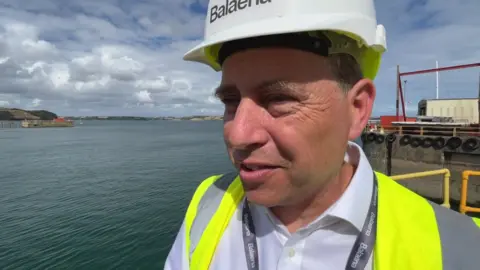 Drystan Jones. He is stood on a dock with the water behind him. He is wearing a high visability vest and a hard hat. He has short grey hair and a is wearing a white shirt.