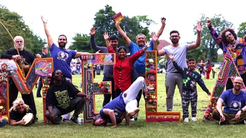 Middlesbrough Mela A group of people using celebratory gestures in front of waist-high letters spelling MELA