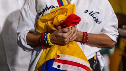 A woman holds Venezuelan flag clutched to her chest. She is wearing multiple bracelets, including a red one, blue one and a yellow one - the colours of the country's flag. 