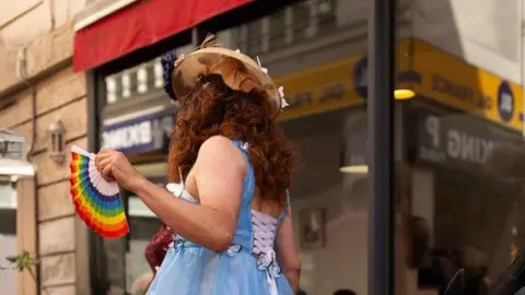 A drag performer is in a street. They're wearing a curly auburn wig and a blue dress, that is laced up at the back and holding a rainbow coloured fan. 
