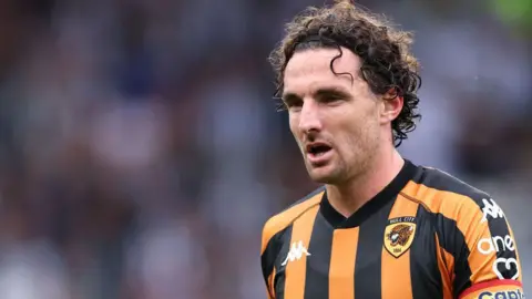 Robbie Jay Barratt - AMA/Getty Images Lewie Coyle playing football for Hull City. He is wearing a traditional black and amber striped football shirt featuring the club's Tiger crest. He has long curly brown hair and is also wearing a rainbow-striped captain's armband.