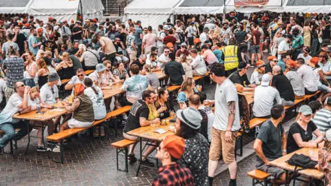 Hundreds of people wearing summer clothes are sat on benches, laughing and sipping beer from glasses. There are white gazebos in the background where people are queuing.