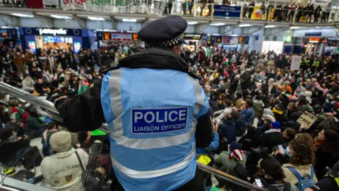 Getty Images A police liaison officer stands overlooking a large crowd of protesters gathered inside a busy London train station, with banners and Palestinian flags visible in the background.