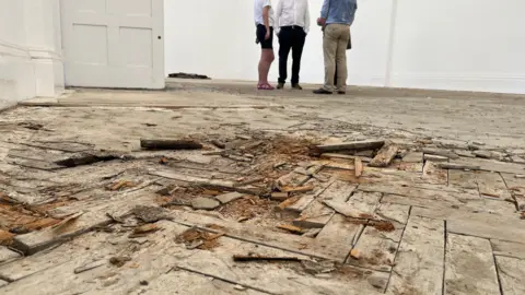 Wooden floorboards, which are broken up, in the Mill Road library building. People are standing in the distance.