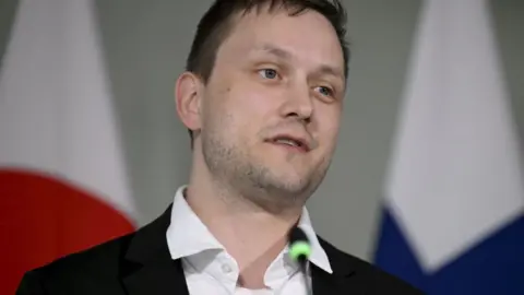 Roni Rekomaa/Reuters Jens-Frederik Nielsen, a man with short brown hair, is wearing a black blazer and white shirt and is stood in front of two flags
