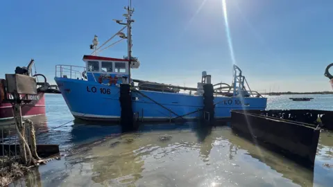 John Fairhall/BBC A blue boat with a tall mast sits in a body of water. The water is shining and the sky above is blue with some lens flare.
