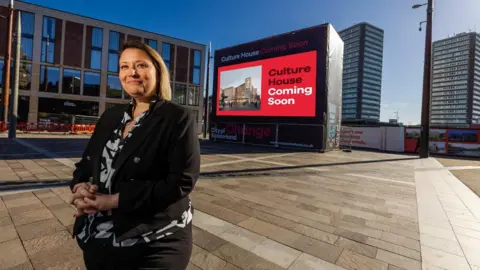 Leanne Littlewood, in a black suit and black and white geometrical shirt, smiles at the camera at Keel Square. She is standing near to where Culture House will be. A poster in the background reads Culture House Coming Soon.