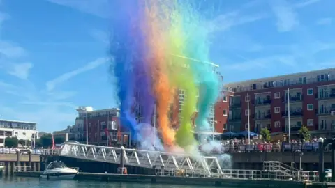 Laura Jackson Colourful smoke rises up towards the sky. People are gathered on the dockside, with blocks of flats behind them.