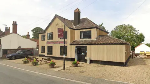 A two‑storey village pub clad in pale vertical timber, with a tiled roof and central chimney, facing a quiet road. A gravel forecourt contains wooden picnic tables and potted plants. Adjacent cottages and a small car park sit to one side, with trees and fencing behind.
