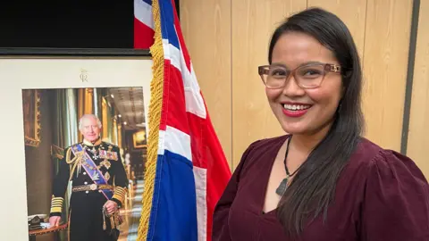 BBC An Asian woman wearing glasses and a burgundy dress stands next to a picture of the King which is draped by a Union Jack