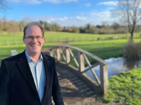 Labour Party Labour candidate Tim Starkey in a dark suit and shirt, with a bridge and field in the background