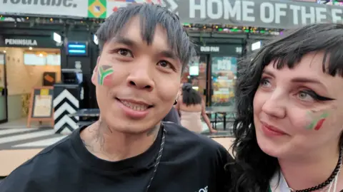 A man and a woman stand in Croydon's Box Park, each with a South African flag painted on their cheek.