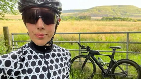 Sara Carter Morgan A woman in large cycling glasses and a black cycling helmet wearing a black cycling jersey with white circles, stood in front of a black bicycle in front of a silver fence. Behind the fence is a field and a hill in the background