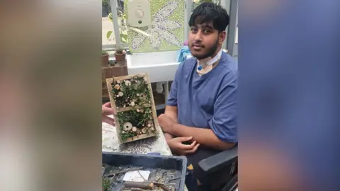 15-year-old Ayuub sits in a wheelchair at a table, his arms are crossed. Someone holds a craft project in front of him. He has a tracheostomy tube in his throat.