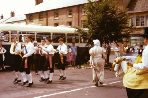 Ray Stokes Morris dancers in traditional outfits in front of an old bus, and also someone in a horse outfit, with people watching in the background.