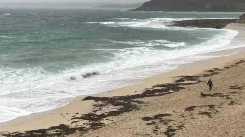 A person and their dog walk along a beach in Cornwall with the stormy sea to their right.