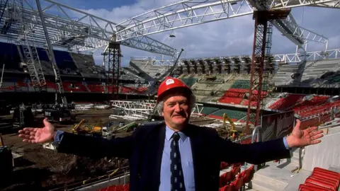 Getty Images Glanmor Griffiths in a red hard hat, light blue shirt, dark jacket and WRU tie. He faces the camera, in the background are cranes and diggers during the early stages of the Millennium Stadium's construction 