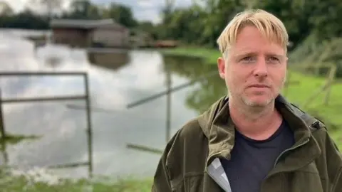 Andrew Davis stands in front of floodwater on his farm. He has short blonde hair and is wearing a dark green jacket.