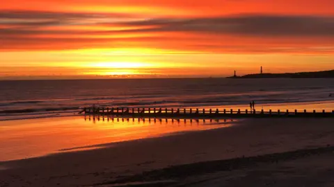 Dennis Grattan A vivid red and orange sunrise spreads across the horizon above a sandy beach. Reflections of the colourful sky shimmer on the wet sand, while a groyne and distant lighthouse stand in silhouette against the sea.