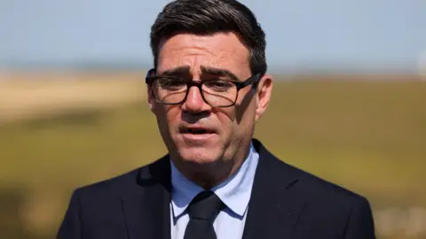 Mid shot of Andy Burnham wearing a black suit and black tie with black-framed glasses. In the distance is the white cliffs of Dover.
