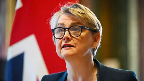Leon Neal via Getty Images UK Foreign Secretary Yvette Cooper wearing black rimmed glasses and a blue blazer, sitting in front of the British flag.