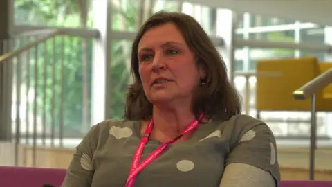 Joss Wills is sitting in a waiting area inside County Hall in Truro, Cornwall Council's headquarters. She is a middle-aged woman with shoulder-length brown hair and wearing a grey-green top with white polka dots. She also has a pink lanyard around her neck. 