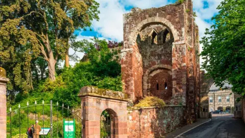 Getty Images Entrance to Rougemount Castle, Exeter, Devon, England. It is a large ruin of a red stone structure with a newer black gate on the left side.