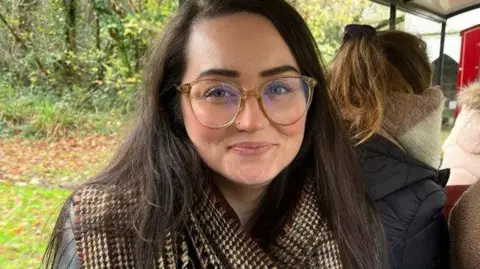 Hannah-Abby Hannah-Abby, a 35 year old woman with brown hair and blue eyes. She is wearing glasses with pale beige frames and is sitting on a bus going through a rural area