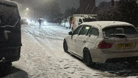 Liv A white car on a road covered in snow. There are other cars on either side of the road. 