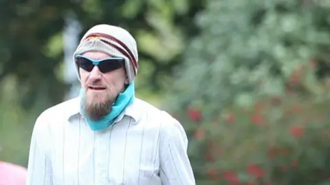 A man wearing a striped shirt and and hat while carrying sunglasses.