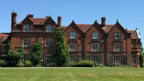 A large brick-built three-storey college building. There are trees and bushes at the foot of the building and in the foreground is a large lawn.