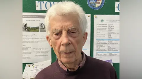 Peter McNamara stands in front of a pin board in a care home. There are details from the council on flyers. Peter is an elderly man with wispy silver hair and a maroon coloured jumper with a red shirt underneath. He is not smiling.