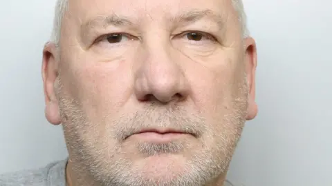 A custody picture of a man with short grey hair and a beard. He is wearing a grey T-shirt.