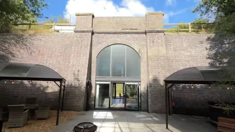 A grey brick railway arch, which is filled by large glass panes. At the front is a patio and on top is a road with a car travelling over it.