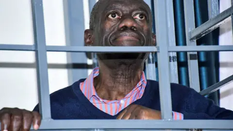 Reuters Ugandan opposition leader Kizza Besigye in a stripped orange and purple shirt and navy jumper seen behind bars in a steel dock at a military court in Kampala - December 2024.